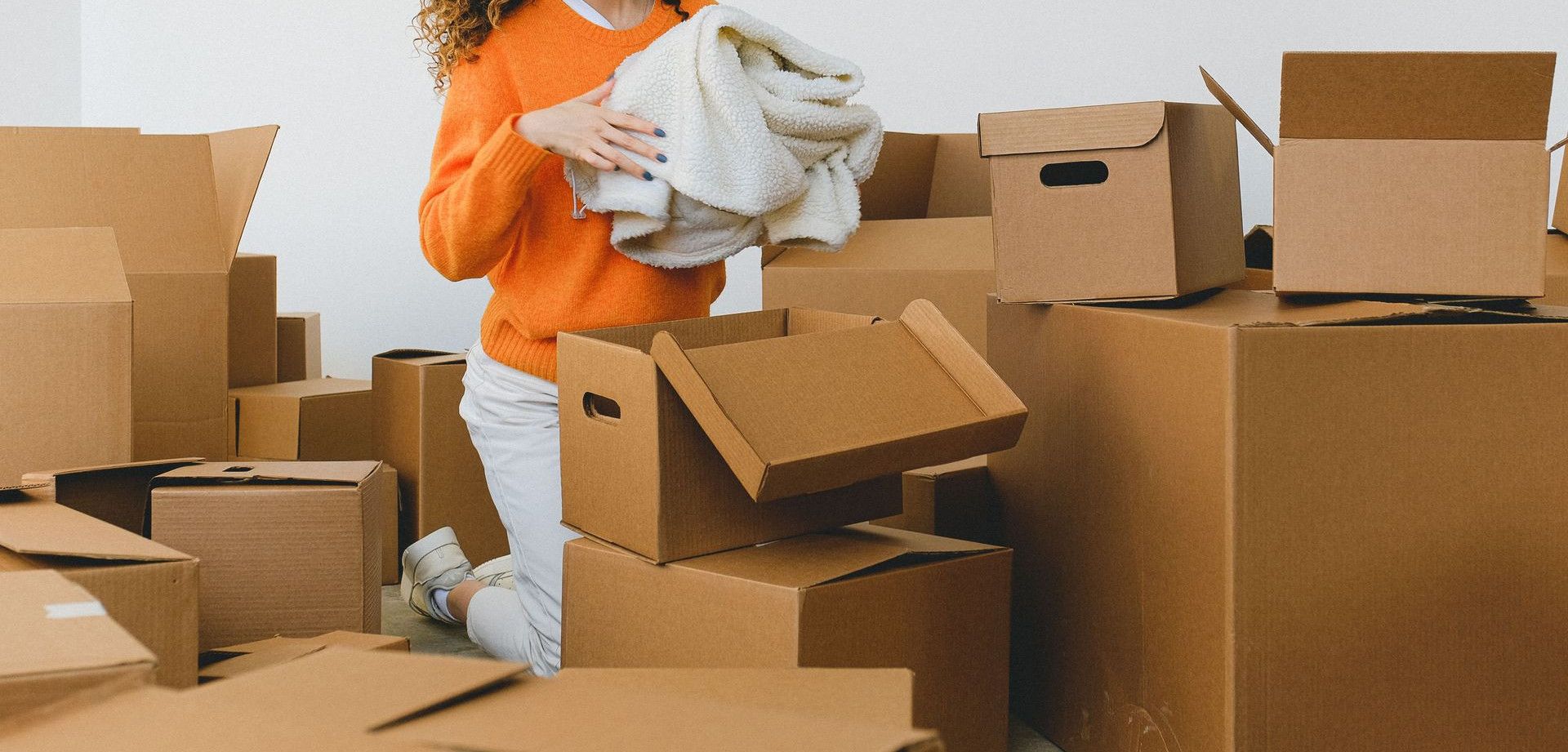 Concentrated young lady with curly hair standing on knees on floor and packing clothes into carton boxes before moving into new flat