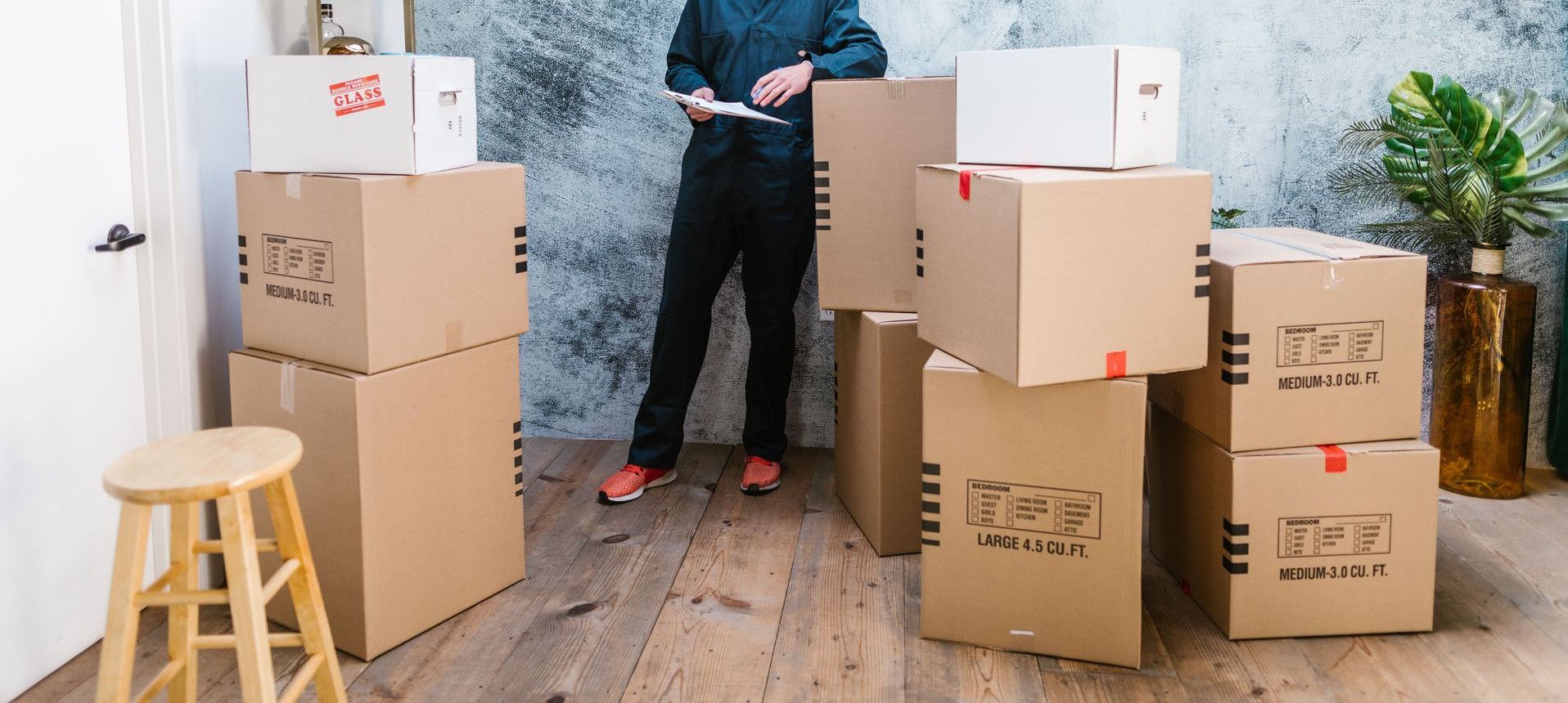 Smiling mover in coveralls standing with parcels, clipboard in hand, indoors.