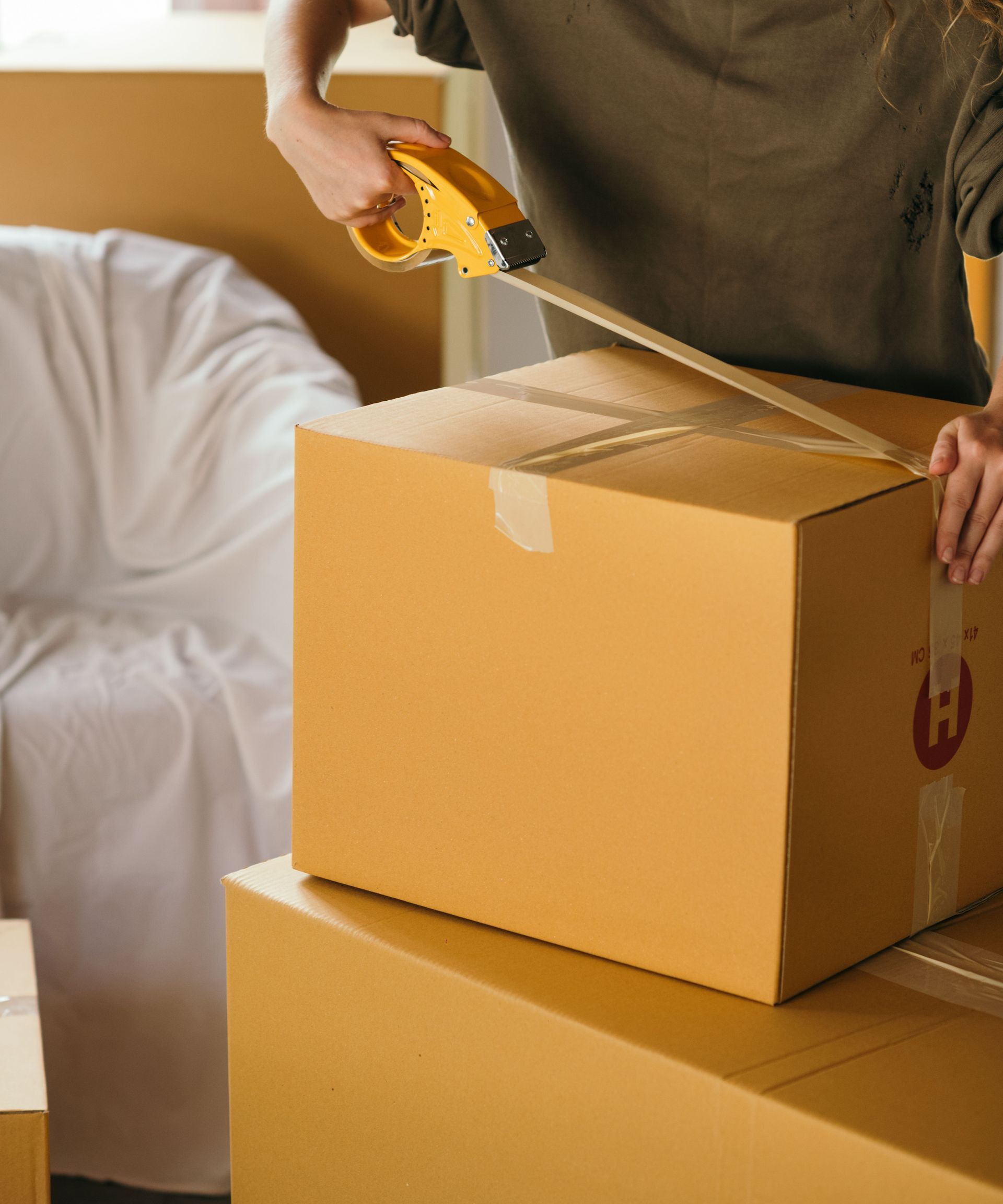 Crop female wearing casual clothes standing while packing carton boxes with scotch tape in modern apartment during preparing for relocation