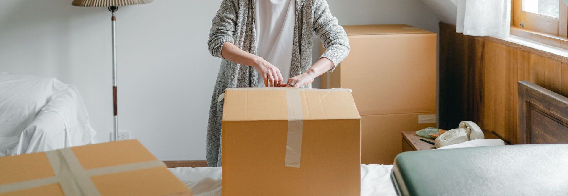 Slim female in cardigan unpacking cardboard box put on bed close to suitcase while standing near window in attic style room in daylight